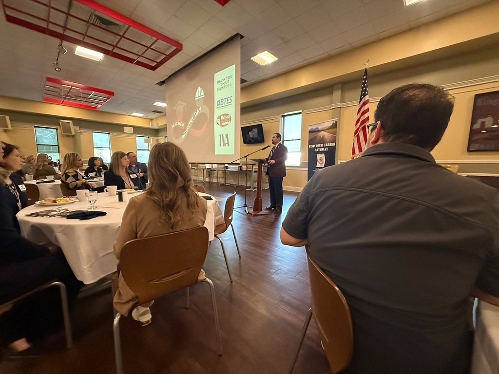 A group of adults sits at round tables during a formal event while a speaker stands at a podium addressing the room. A large screen displays the Teacher Industry Day logo and sponsor logos, and an American flag stands nearby. Attendees listen as the presentation takes place in a banquet-style setting.