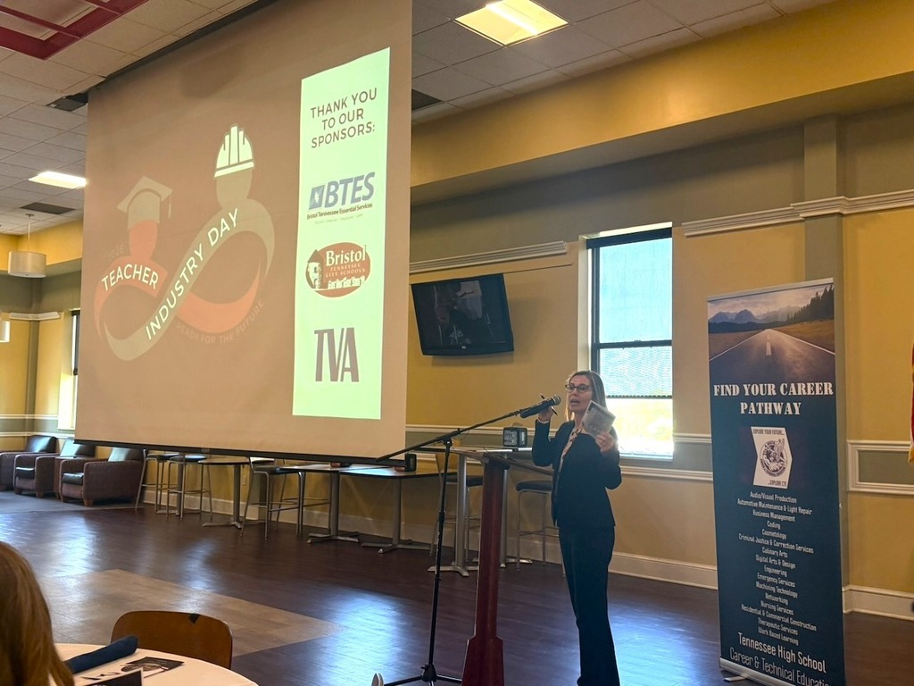 A woman speaks at a podium during Teacher Industry Day, holding notes while addressing the audience. Behind her, a large screen displays the event logo and a “Thank You to Our Sponsors” slide featuring BTES, Bristol Tennessee City Schools, and TVA. A banner to the side reads “Find Your Career Pathway,” highlighting career and technical education pathways.