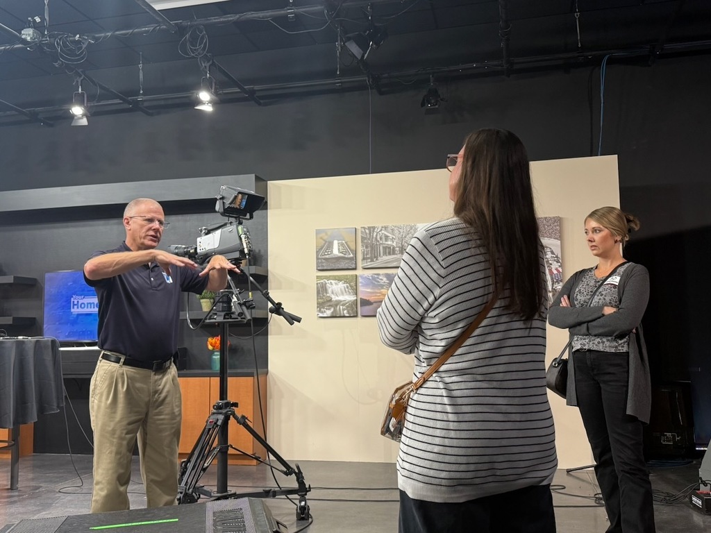 Several adults stand in a television studio with professional cameras, lighting equipment, and set pieces. One person gestures while speaking near a camera on a tripod, as two others listen nearby. The studio includes overhead lighting rigs, a monitor, and staged backdrops, showing a behind-the-scenes look at video production.