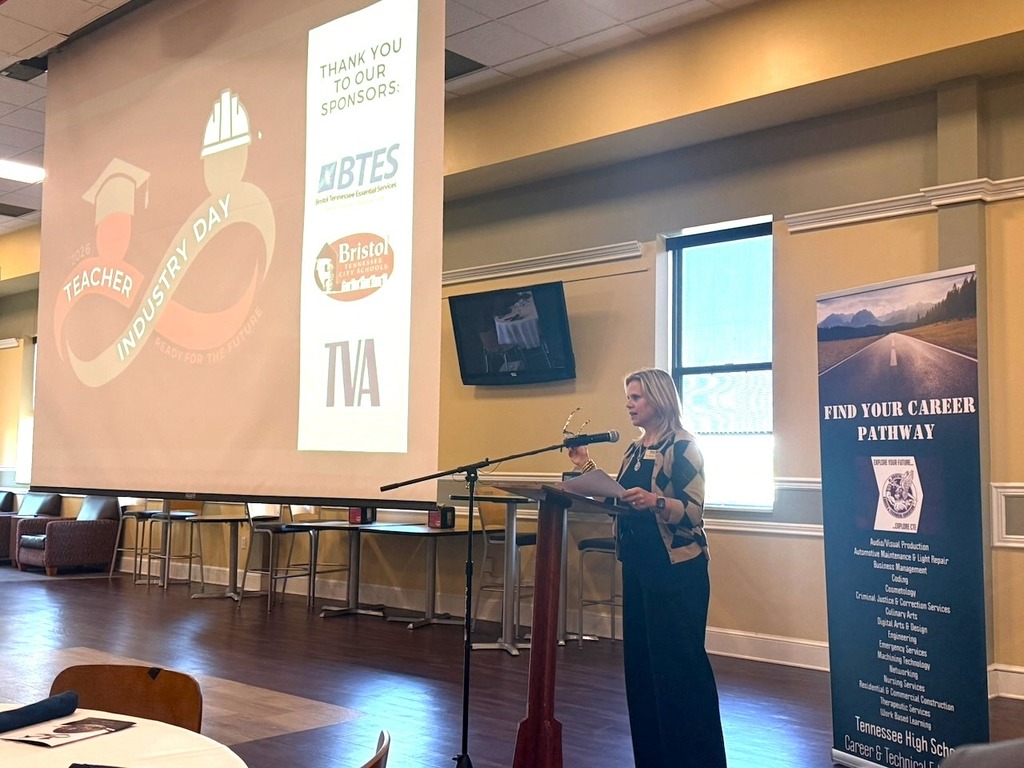 A woman stands at a podium speaking into a microphone during Teacher Industry Day in a large meeting room. Behind her, a projected screen displays the event logo and a “Thank You to Our Sponsors” slide featuring BTES, Bristol Tennessee City Schools, and TVA logos. A banner to the right reads “Find Your Career Pathway” and lists various career fields. Tables and chairs are set up throughout the room.