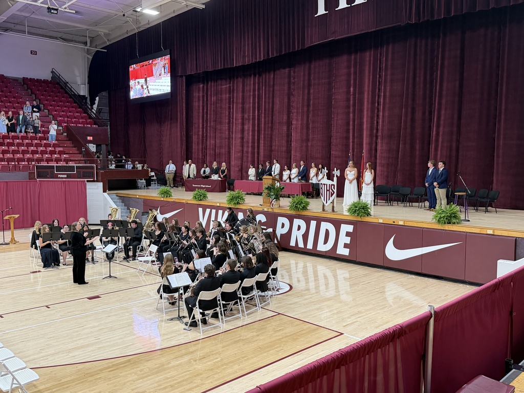 Wide view of Tennessee High School’s Class Night ceremony inside Viking Hall. The school band performs on the gym floor while students and speakers stand on stage in formal attire beside the BTHS shield and ceremonial axes. Maroon curtains frame the stage, and “VIKING PRIDE” is displayed prominently along the front wall of the gym. Spectators are seated in the bleachers as the ceremony takes place.