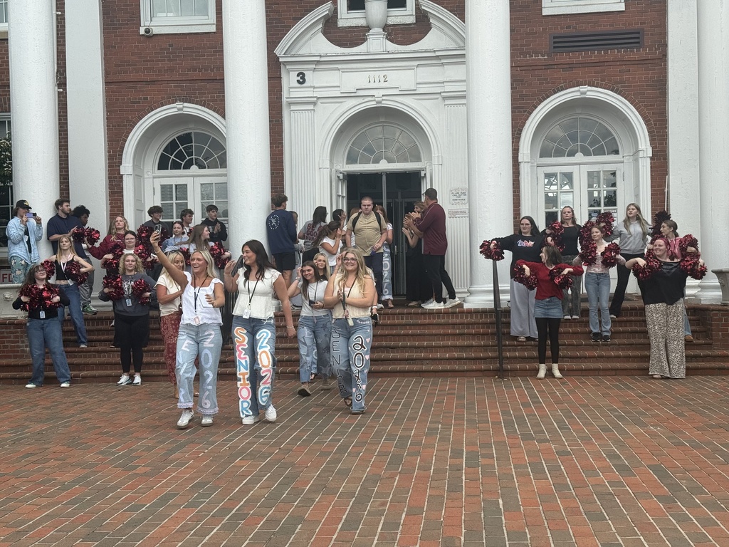 Students and staff gather on the front steps of Tennessee High School during a celebratory send-off. Several seniors wearing decorated jeans with “Senior 2026” painted on the legs walk across the brick courtyard while cheerleaders hold maroon pom-poms on either side of the entrance.