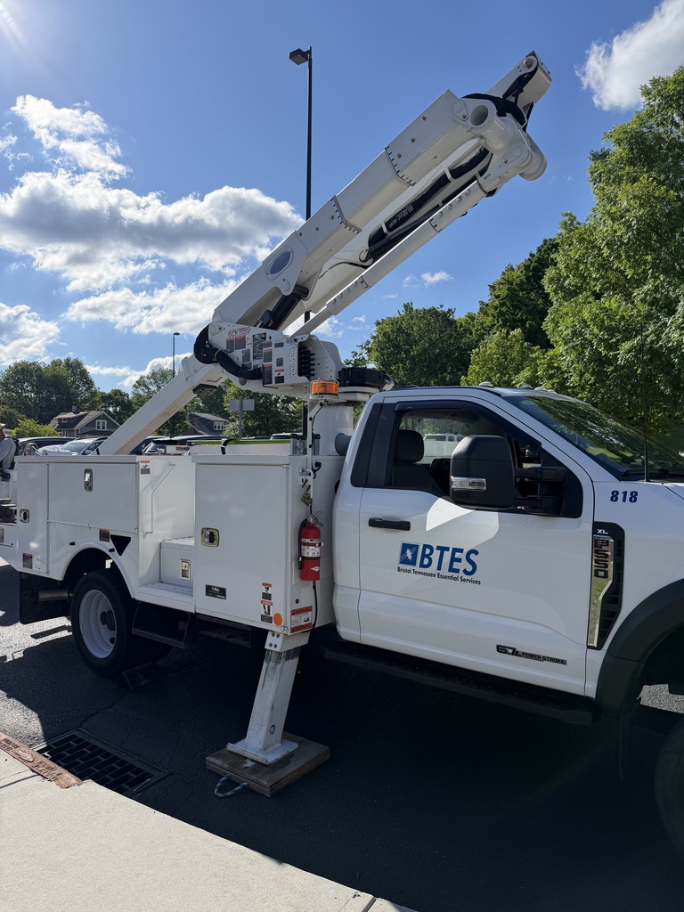 A BTES bucket truck is parked outside on a sunny day with its boom arm extended high into the air. The white utility truck, labeled “BTES Bristol Tennessee Essential Services,” is set up for the school’s Egg Drop Challenge with trees and a bright blue sky in the background.