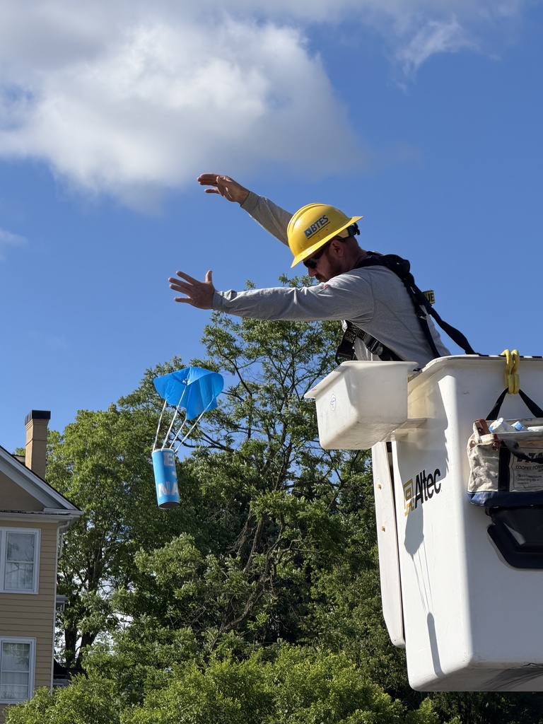 A BTES worker wearing a yellow hard hat and safety harness releases a student-designed egg drop container from a bucket truck during an outdoor STEM activity. The blue parachute-topped container floats downward against a bright blue sky with clouds and trees in the background.