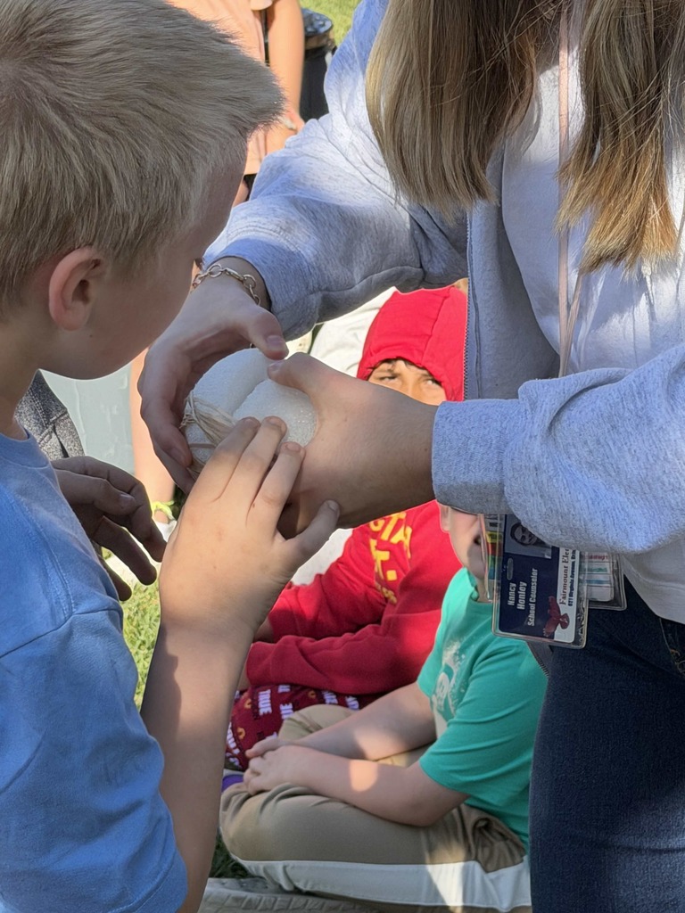 Close-up of a student carefully holding an egg-drop project wrapped in protective material while another student and classmates gather nearby outdoors during the school’s Egg Drop Challenge activity.
