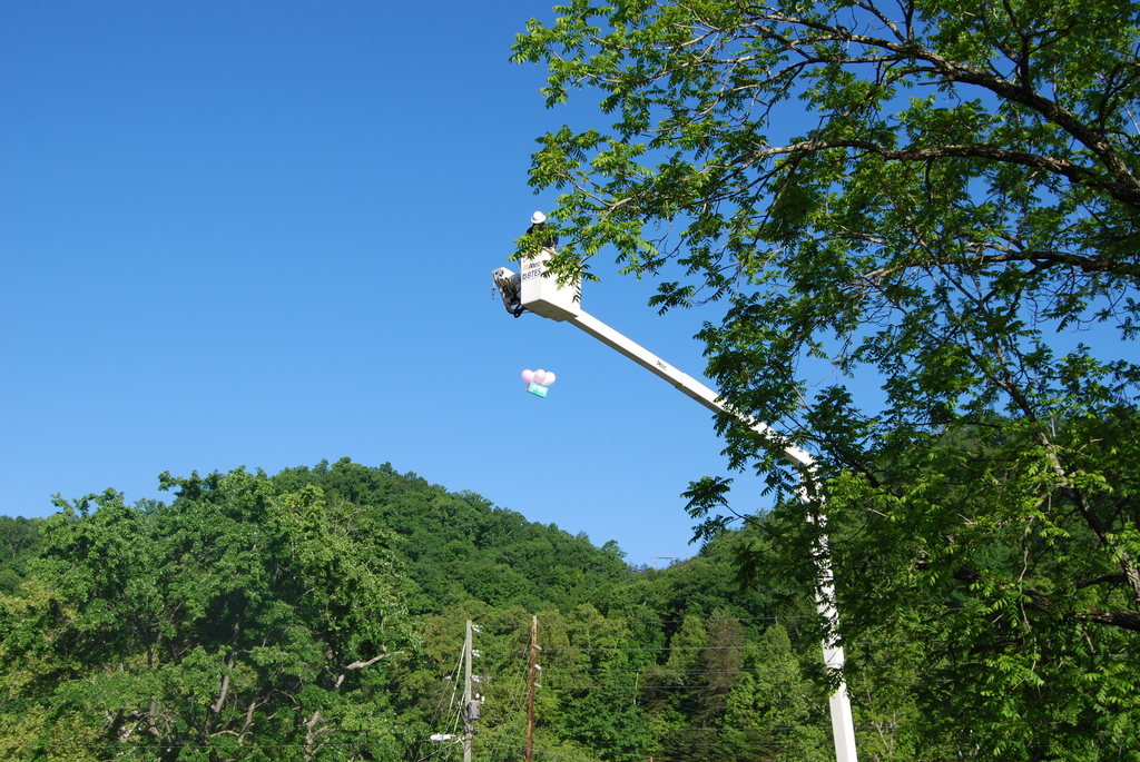 A BTES utility worker stands in a raised bucket truck high above the ground beside tree branches as a small egg-drop container with balloons attached falls through the air against a bright blue sky and green hillside backdrop.