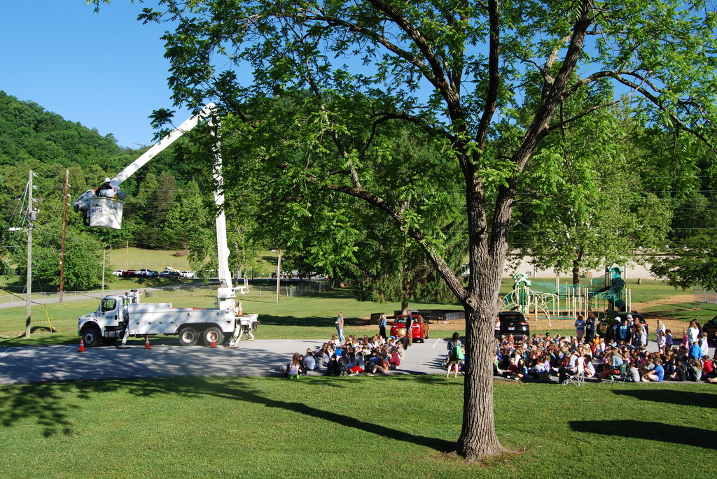 Wide view of an outdoor school event with dozens of elementary students seated on pavement and grass near a playground while a BTES bucket truck extends high into a large tree. Two utility workers sit in the raised bucket as students watch the demonstration on a sunny day surrounded by green hills and trees.