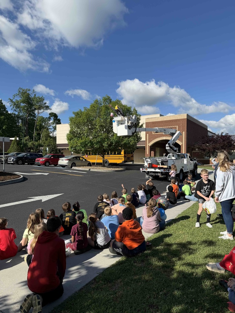 Students sit along a sidewalk outside the school watching an Egg Drop Challenge demonstration as a utility worker in a raised bucket truck prepares to drop a student-created project from above. A school bus, parked cars, and the school building are visible in the background under a bright blue sky with clouds.