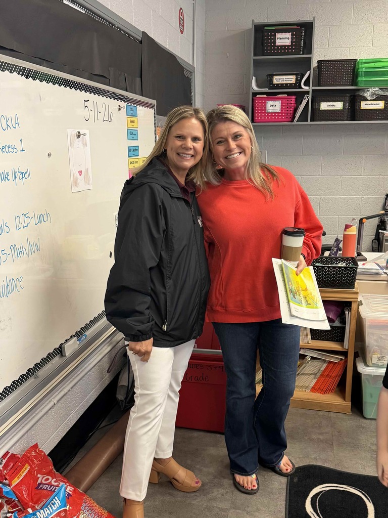 Two smiling women stand together in an elementary classroom beside a whiteboard. One woman wears a black jacket with white pants, and the other wears a bright red sweatshirt and jeans while holding a coffee cup and papers. Classroom shelves with labeled bins, teaching materials, and student supplies are visible in the background.