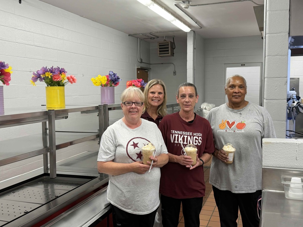 Four Bristol Tennessee City Schools staff members stand together in a school cafeteria kitchen holding iced coffee drinks topped with whipped cream. Stainless steel serving counters line the room, and colorful flower arrangements in painted cans decorate the shelves behind them. One staff member wears a Tennessee High Vikings shirt, while another wears a “peace, love & lunch” nutrition services shirt.