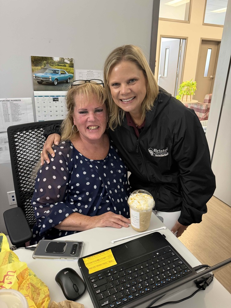 Two smiling women pose together in an office workspace. One woman is seated at a desk wearing a navy blue polka-dot blouse, while the other stands beside her in a black Bristol Tennessee City Schools jacket. A large iced coffee drink, computer keyboard, mouse, and phone are on the desk, with a calendar and framed car photo visible on the wall behind them.