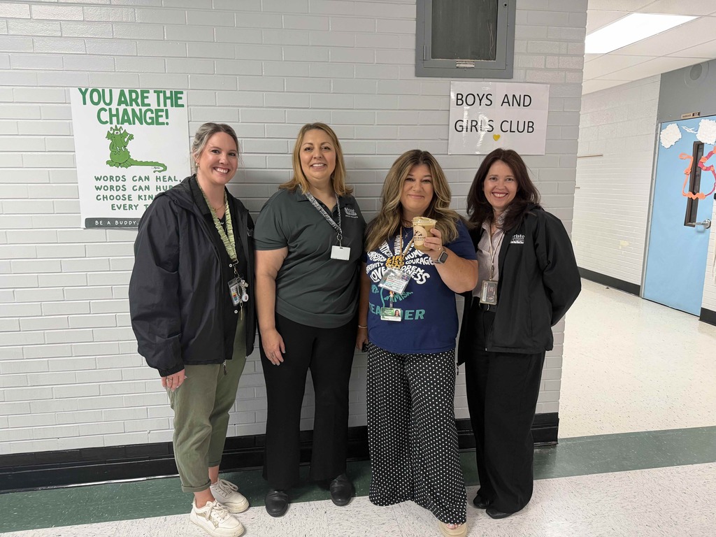 Four Bristol Tennessee City Schools staff members stand smiling together in a school hallway beneath posters promoting kindness and the Boys and Girls Club. One staff member holds an iced coffee drink. The group is dressed in casual professional attire with staff badges visible. White brick walls and colorful classroom decorations are visible in the background.