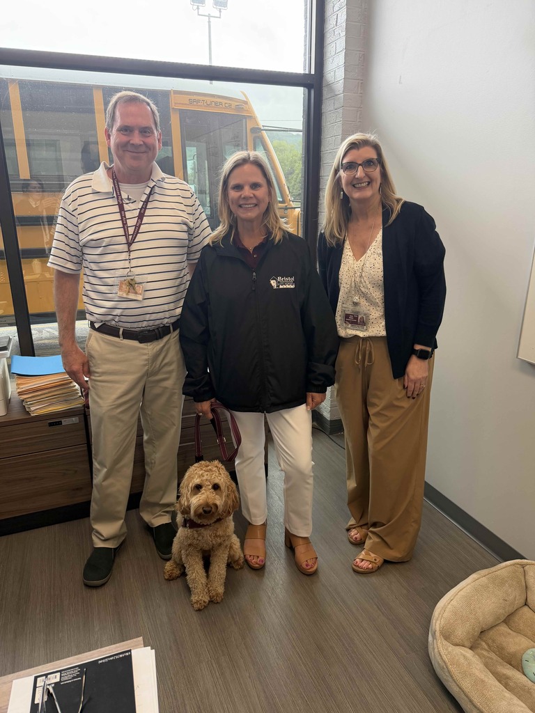 Three Bristol Tennessee City Schools staff members stand together in an office area beside a curly-haired therapy dog. A school bus is visible through the large window behind them. One staff member wears a BTCS jacket, while the others are dressed in business-casual attire with staff badges. A dog bed sits on the floor nearby.