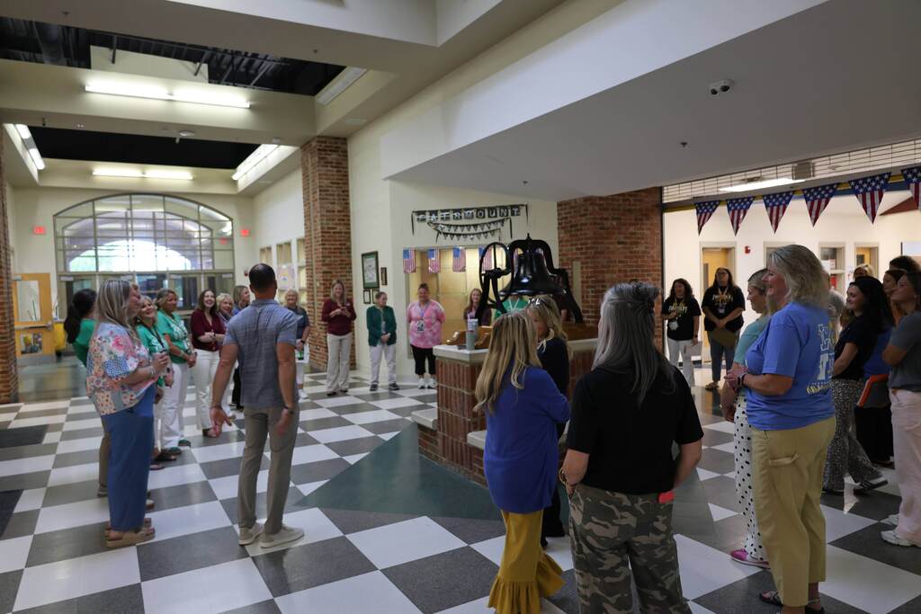 A group of educators and staff members stand in a large circle inside a school commons area, participating in a team-building or discussion activity. The space features a black-and-white checkered floor, exposed brick columns, large windows, and a historic bell display at the center. Patriotic bunting hangs along an upper walkway in the background as participants listen and engage with one another in a bright, open setting.