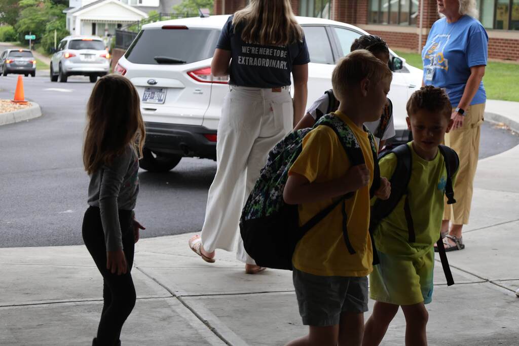 Several elementary-age students wearing backpacks arrive at a school entrance as staff members greet them during morning drop-off. Two children walk together toward the building while another stands nearby, and adults supervise and welcome students at the curb. Vehicles are visible in the drop-off lane outside the school, creating a busy and welcoming start to the day.