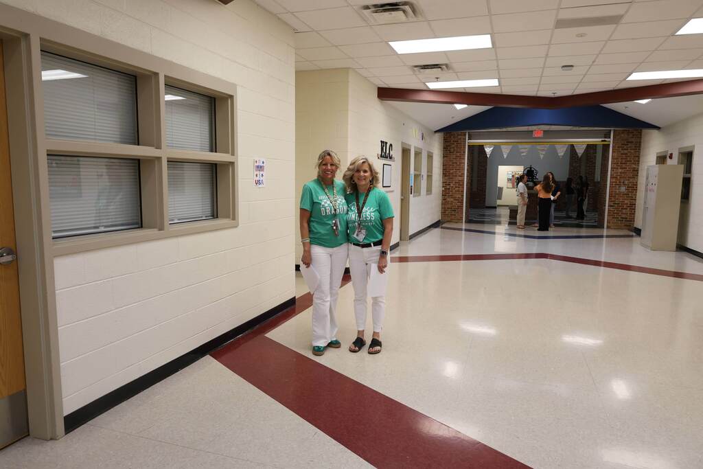 Two school staff members stand together smiling in a bright school hallway. Both are wearing matching green school-themed T-shirts and white pants, with staff identification badges visible. In the background, the hallway opens into a larger common area where several people are gathered and talking near exposed brick columns and decorative bunting. The clean, well-lit school interior features classroom windows, neutral walls, and polished floors.