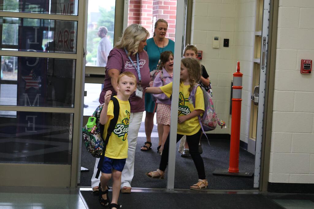 A school staff member walks hand in hand with two young students wearing bright yellow Summer STREAM Camp T-shirts and backpacks as they enter the school. Other children and adults follow behind them near the entrance. A colorful backpack with a unicorn design is visible in the foreground, capturing the excitement and energy of students arriving for the start of camp.