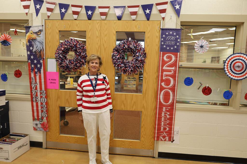 A school staff member stands smiling in front of library doors decorated with patriotic red, white, and blue wreaths and banners. The display celebrates America’s 250th anniversary, featuring “1776–2026” and “250 Freedom” banners, patriotic bunting, and festive paper decorations. The staff member is wearing a red-and-white striped shirt with blue stars and white pants, matching the patriotic theme of the library entrance.