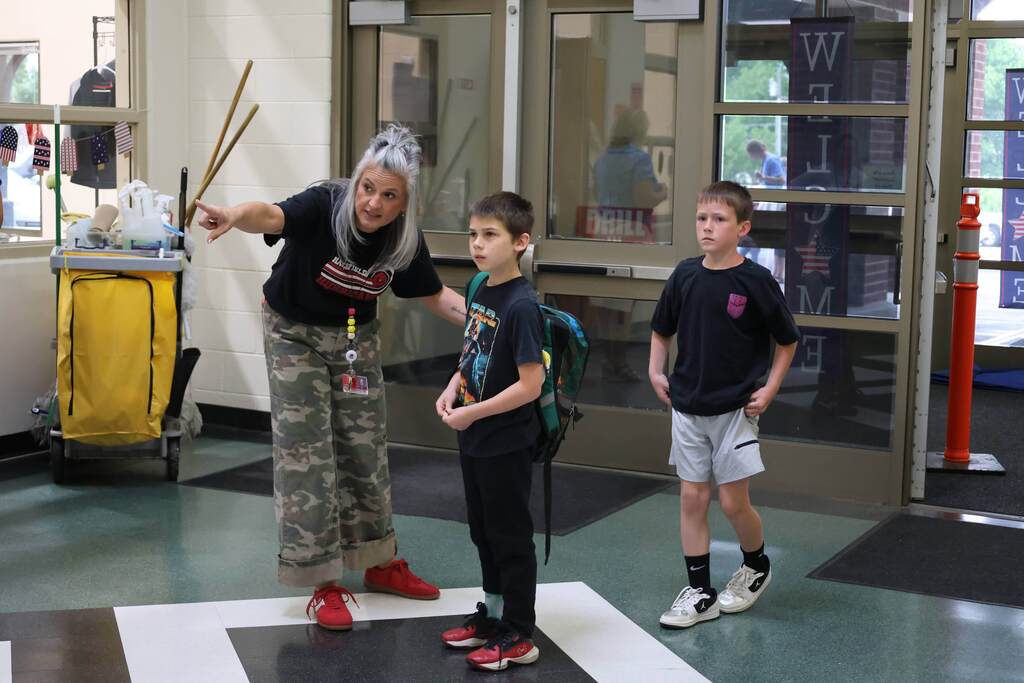 A school staff member kneels slightly and points toward a destination while helping two elementary-age students find their way inside the school. The students, both wearing backpacks, listen attentively near the entrance as they prepare to head to their classrooms or camp activities. Patriotic decorations and welcome signs are visible in the background, creating a friendly and supportive first-day atmosphere.