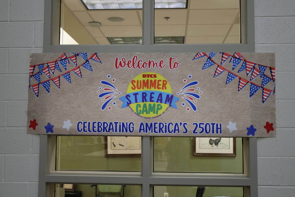 A colorful banner hangs in a school hallway window welcoming students to BTCS Summer STREAM Camp. The banner features patriotic red, white, and blue decorations, including flag bunting, stars, and fireworks graphics, along with the message, “Welcome to BTCS Summer STREAM Camp — Celebrating America’s 250th.” The display reflects the camp’s summer theme and recognition of the nation’s upcoming 250th anniversary.