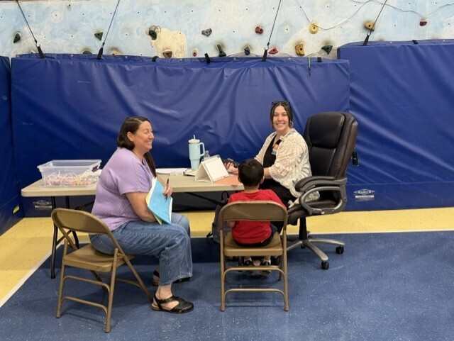 A young child sits with two adults at a registration or screening table set up inside a gymnasium. One adult is seated in a chair reviewing paperwork, while the other sits at a table with a tablet or laptop. Blue wall padding and an indoor climbing wall are visible in the background. The group appears to be engaged in a check-in, assessment, or enrollment activity.