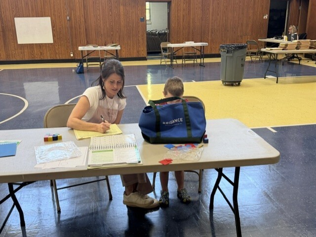 A staff member sits beside a young student at a table in a gymnasium, taking notes while speaking with the child during an assessment or screening activity. Educational materials, manipulatives, and paperwork are spread across the table. Additional screening stations and staff members can be seen in the background.