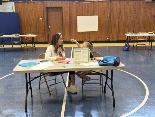 A staff member and a young child sit across from one another at a table in a gymnasium during an assessment or screening activity. The staff member appears to be asking a question while the child gestures in response. Assessment materials, folders, and supplies are spread across the table, with additional screening stations visible in the background.