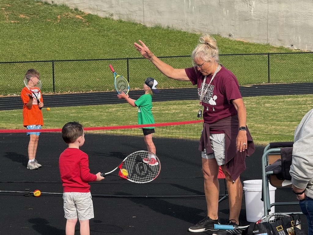 A female instructor wearing a maroon Viking Tennis Camp shirt demonstrates a tennis swing for a young child in a red shirt holding a tennis racket on an outdoor court. Two other young children holding rackets practice in the background.