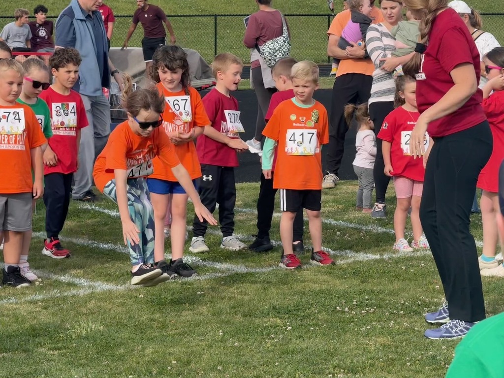 A group of young children wearing numbered race bibs and Power of Play Youth Sports Day t-shirts prepare to compete in a standing broad jump on a grass field. An adult in a maroon shirt observes from the right side.