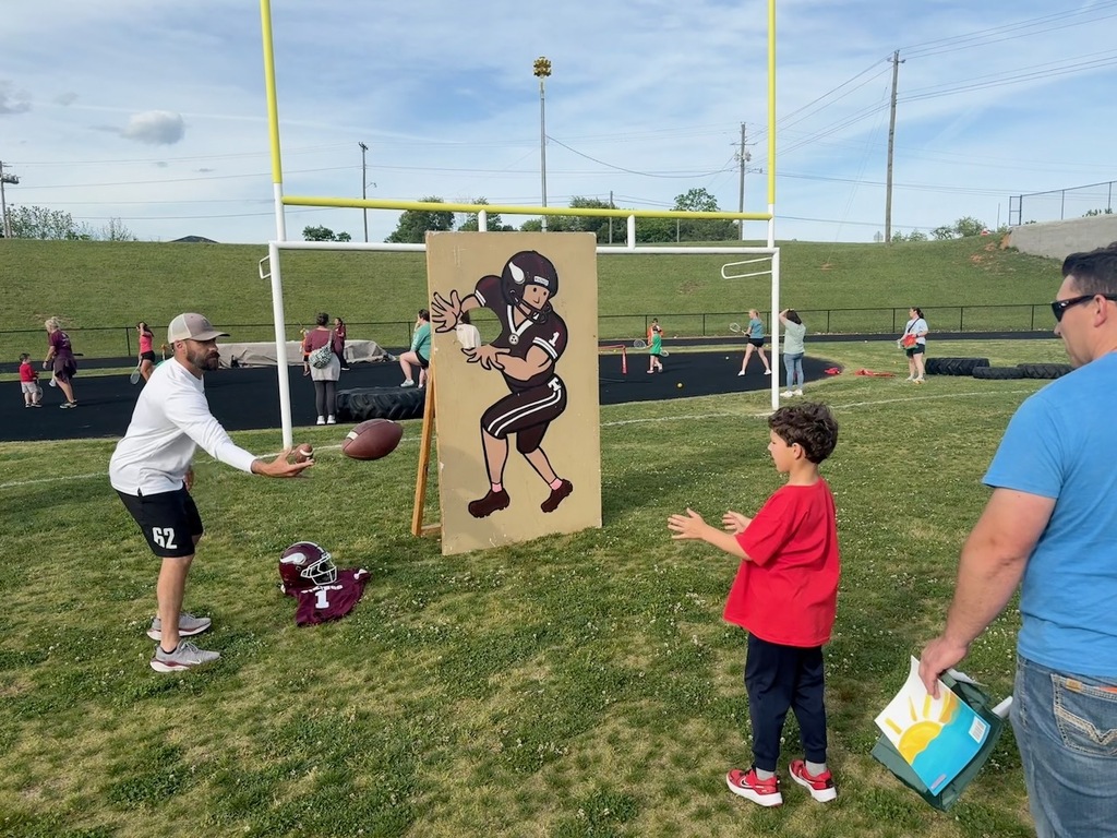 A man in a white long-sleeve shirt tosses a football to a child in a red t-shirt standing in front of a cardboard cutout of a Viking football player. A yellow goal post and a Tennessee High football helmet are visible in the background, with other event activities taking place on the field.