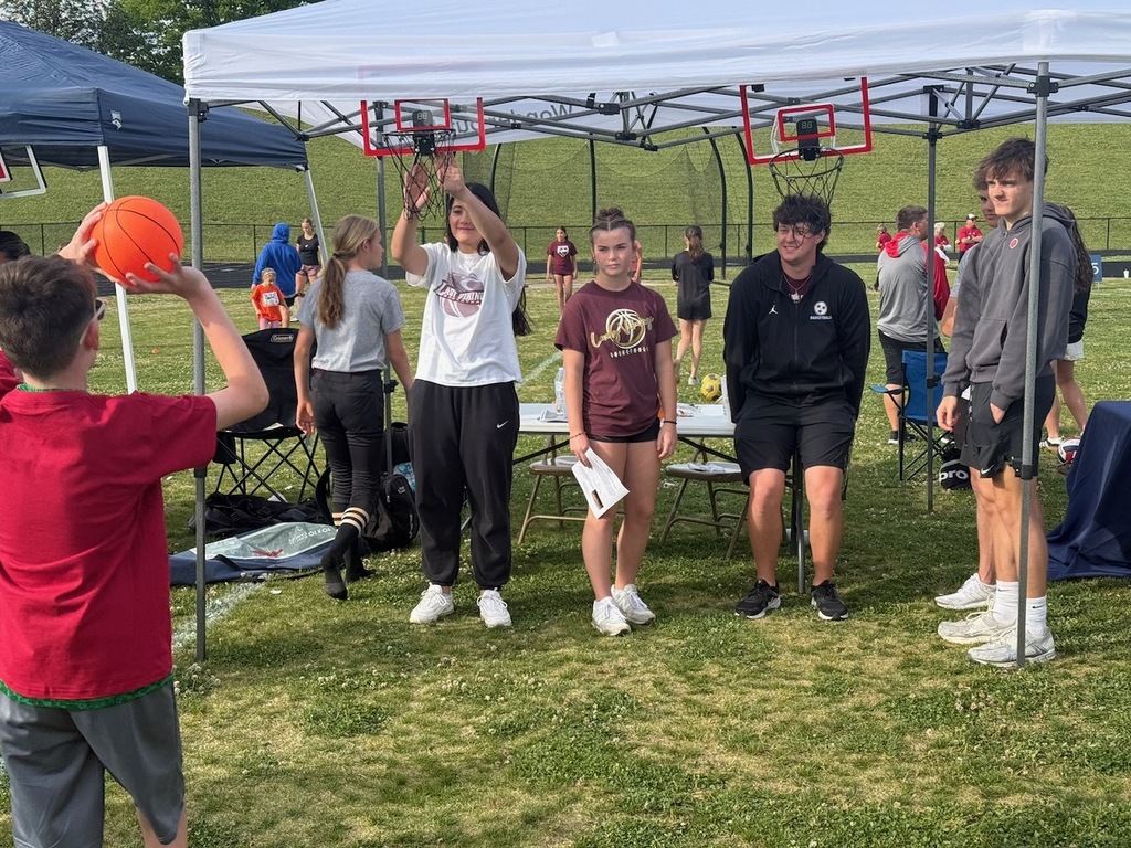 A young child in a red shirt prepares to shoot a small basketball at a portable hoop inside a canopy tent. Several Tennessee High student-athletes in maroon and black athletic wear stand nearby watching, with additional event activities visible on the field in the background.