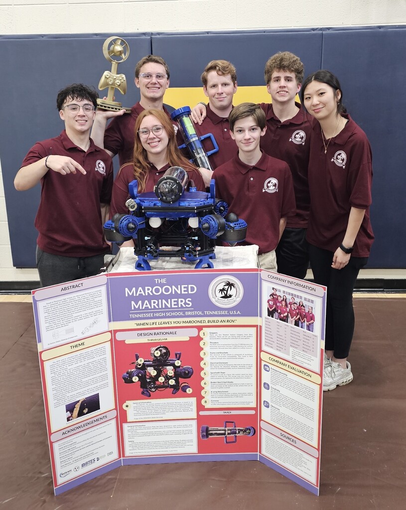 Seven members of Tennessee High School's Viking Robotics team, the Marooned Mariners, stand behind their underwater remotely operated vehicle (ROV) and competition display board. The students wear matching maroon polo shirts and pose with their robot, which is blue and black with multiple thrusters and a mounted camera. One student holds a trophy while another holds a component of the robot. The display board features information about the team's design, company structure, and engineering process. The group is gathered in a school gymnasium after qualifying for the 2026 MATE ROV Underwater Robotics World Championship.