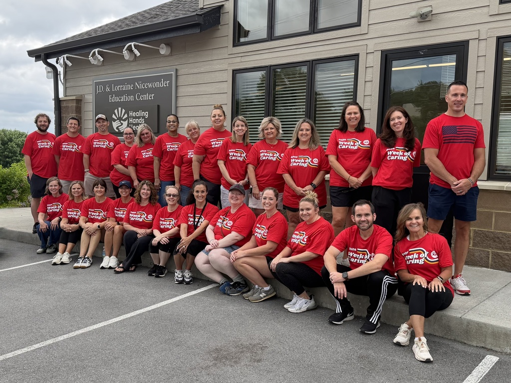 Twenty-six Bristol Tennessee City Schools employees wearing matching red Week of Caring T-shirts pose for a group photo outside the J.D. & Lorraine Nicewonder Education Center at Healing Hands Health. The volunteers are gathered in front of the building's entrance and sign after spending the morning completing service projects as part of United Way of Bristol TN/VA's Week of Caring. The group includes employees standing and seated together on the sidewalk outside the facility.