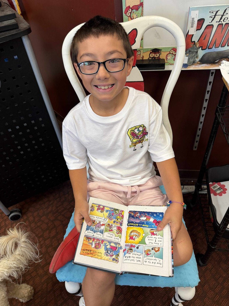 A smiling student sits in a child-sized chair reading a colorful graphic novel during Drop Everything and Read (DEAR) time in the Fairmount Elementary School library.