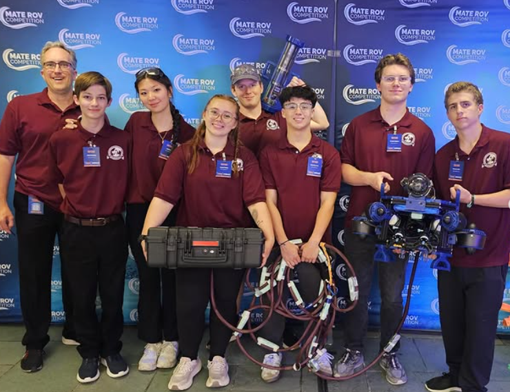 A group of eight Tennessee High School Marooned Mariners robotics team members and their coach pose in front of a Mate ROV Competition backdrop. The students wear matching maroon polo shirts with team logos and event name badges. Several hold underwater robotics equipment, including a large remotely operated vehicle (ROV), coiled tether cables, a rugged equipment case, and a tall blue trophy. The group smiles proudly after competing at the international event.