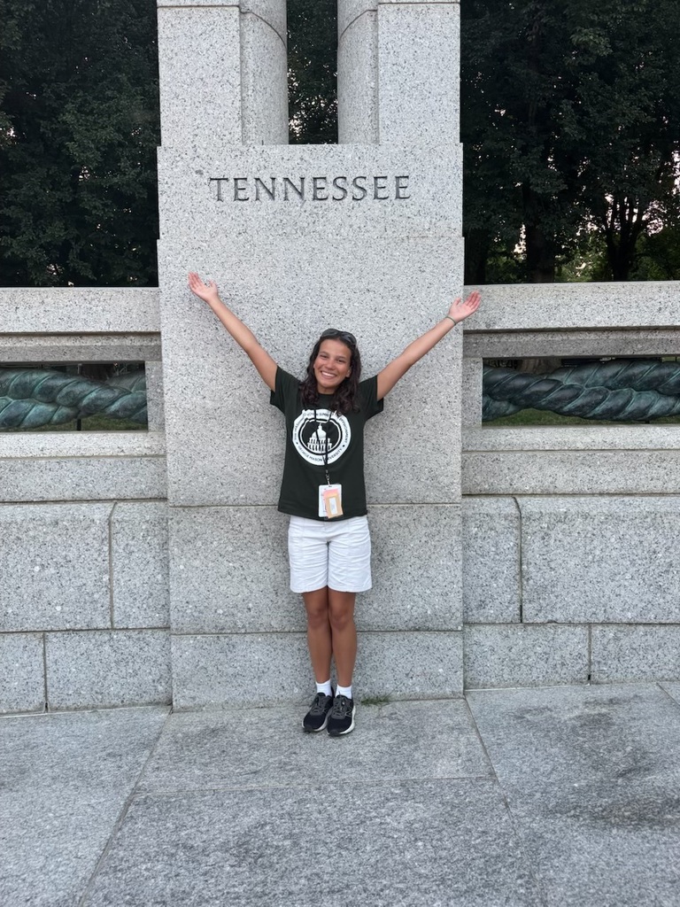 A smiling student stands with both arms raised beneath a large stone monument engraved with the word "TENNESSEE." She is wearing a dark green Washington Youth Summit on the Environment T-shirt, white shorts, gray athletic shoes, and a conference credential on a lanyard. Trees fill the background behind the granite memorial.