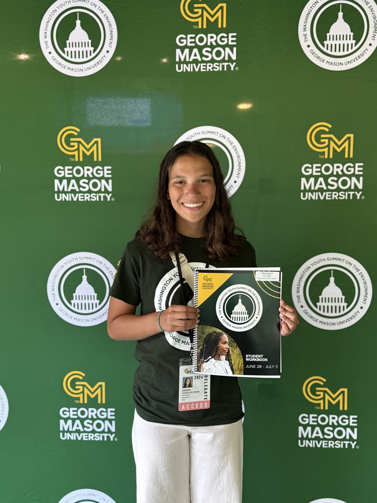 A smiling student stands in front of a green George Mason University backdrop featuring Washington Youth Summit on the Environment logos. She is wearing a dark green summit T-shirt, white pants, and a conference credential. She holds the summit student workbook and poses for a portrait during the conference.