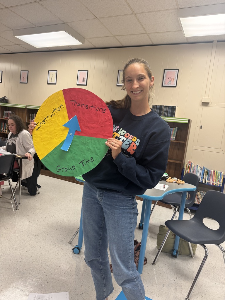 A smiling educator stands in a school library holding a completed classroom routine spinner divided into three color-coded sections labeled "Instruction," "Transitions," and "Group Time," with a blue arrow in the center pointing to one section.