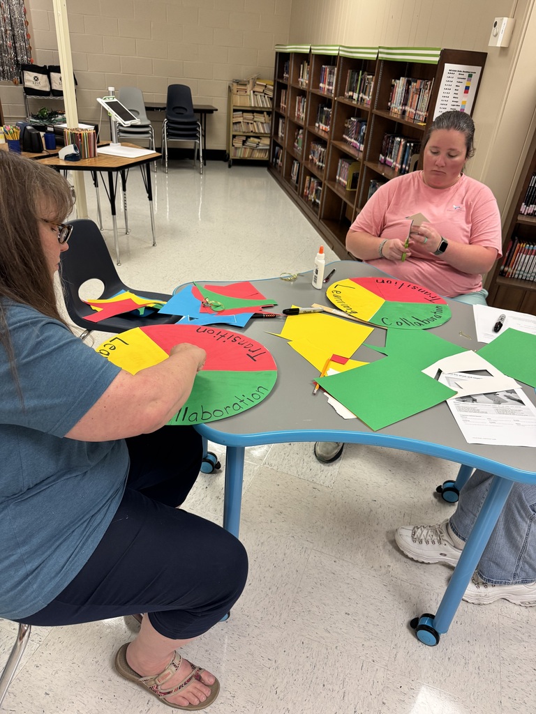 Two educators sit at a table assembling colorful classroom routine spinners made from red, yellow, and green paper. Glue, scissors, markers, and printed instructions are spread across the table as they work.