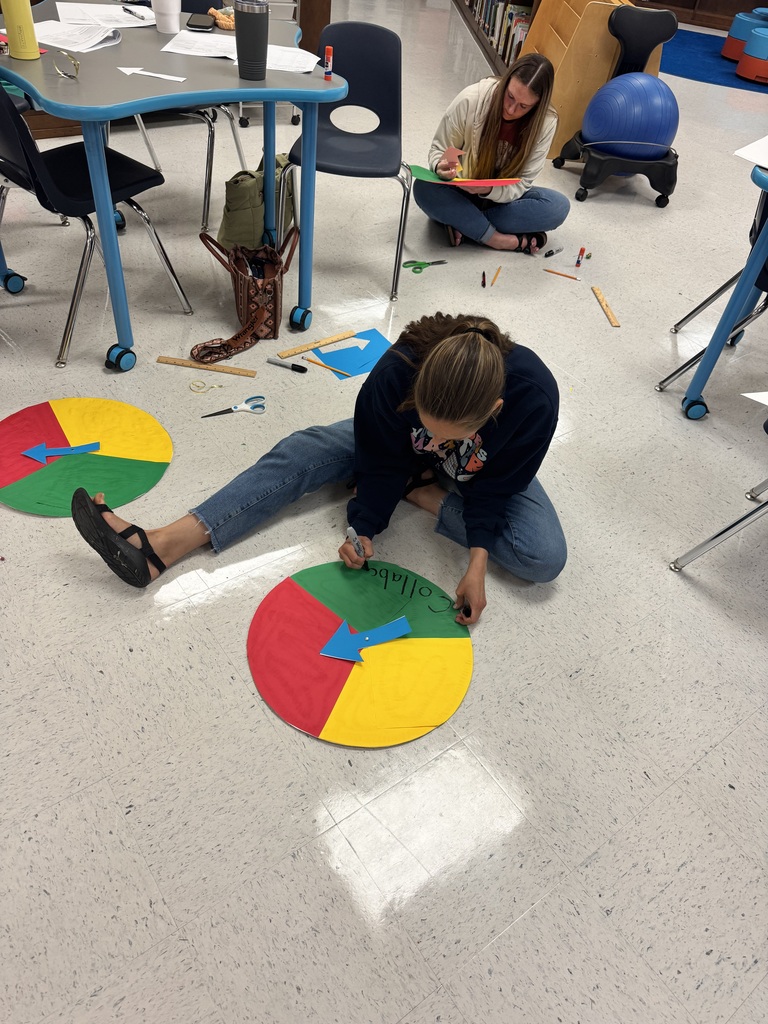 Two educators sit on the library floor writing labels and attaching arrows to completed classroom routine spinners. Craft supplies, rulers, scissors, and paper are scattered around them as they put the finishing touches on their projects.