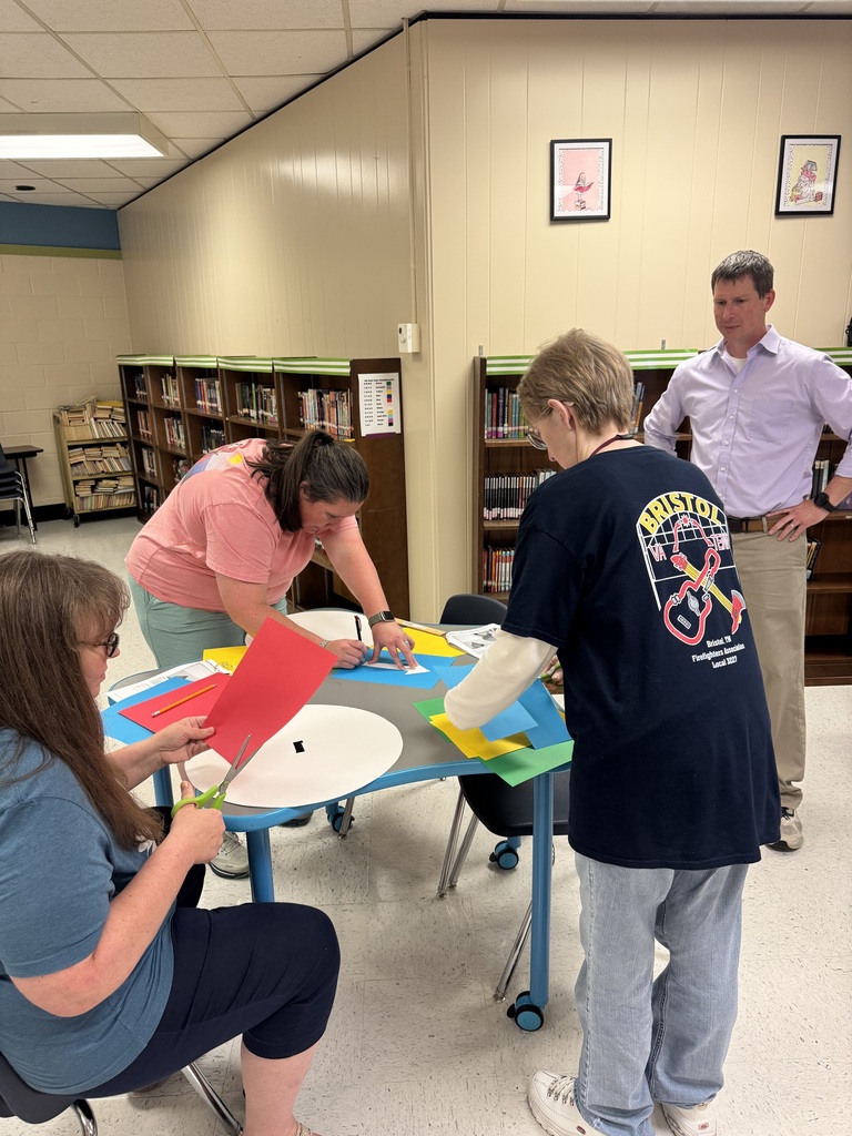 Four educators work together around a table in a school library, cutting colored paper and assembling large circular classroom routine spinners. Another staff member stands nearby observing the activity.