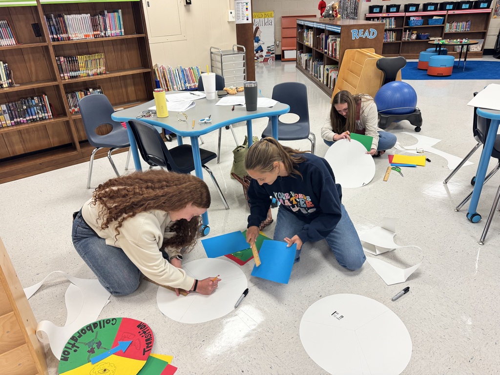 Several educators kneel on the library floor, measuring, cutting, and gluing colored paper onto large circular poster boards as they create classroom routine spinners during a professional learning activity.