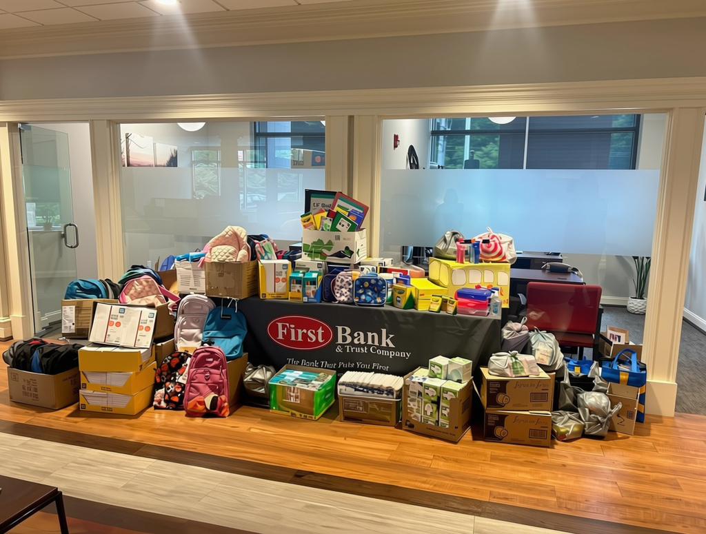 Alt text: A large collection of donated school supplies is displayed on a raised platform inside an office lobby. Backpacks, notebooks, folders, crayons, pencil boxes, tissue boxes, and other classroom necessities are arranged around a table covered with a First Bank & Trust Company banner. The space features wood flooring, glass-walled offices, and bright overhead lighting. Visible text on the banner reads, “First Bank & Trust Company” and “The Bank That Puts You First.”