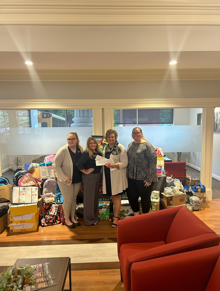 Alt text: Four people stand together indoors in front of a large donation of school supplies and other items. Backpacks, boxes, and educational supplies are arranged around them, and one person holds a check. The group is gathered in a lobby or office area with large windows, wood flooring, and a red armchair in the foreground. A school bus is visible through the windows in the background.