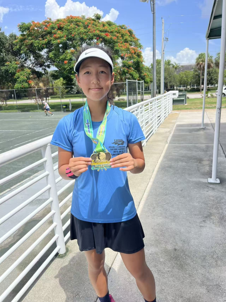 A student stands on an outdoor tennis court holding a medal. She wears a white visor, light blue shirt, and dark skirt. A large tree with orange flowers, a blue sky, and tennis courts are visible in the background.
