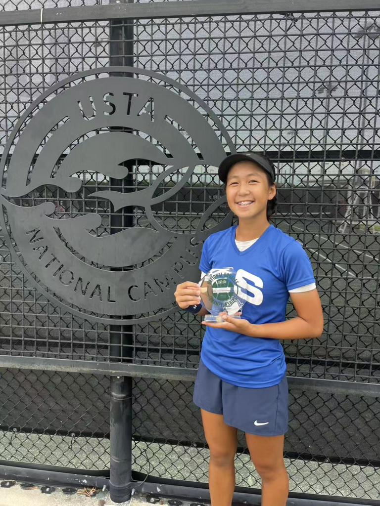 A smiling student wearing a blue shirt and dark athletic shorts stands in front of a large USTA National Campus sign, holding a tennis award.