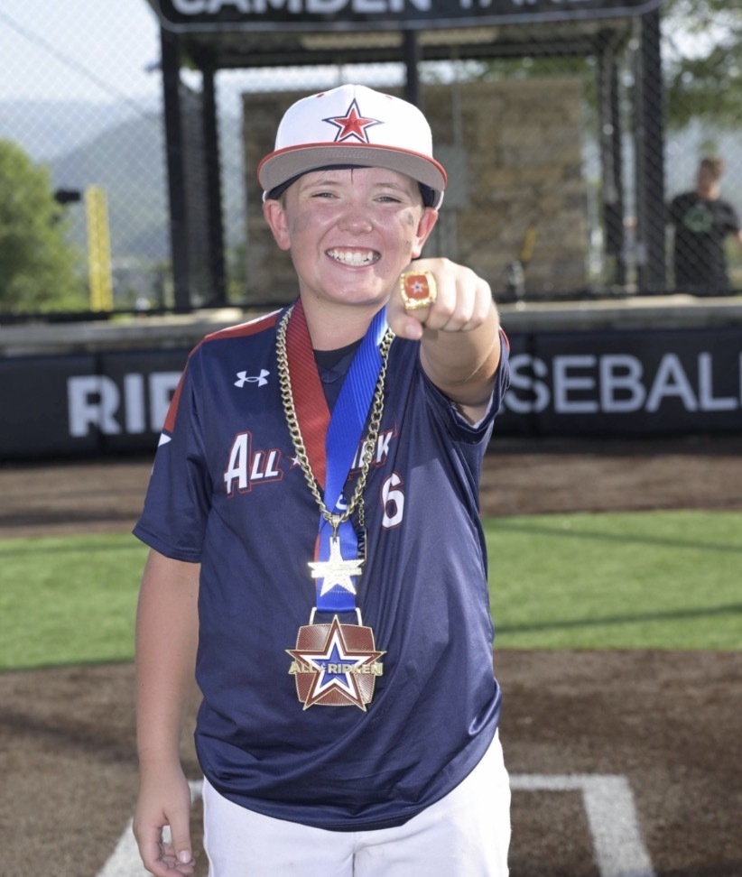 A smiling young baseball player wearing a navy, red, and white uniform and a white baseball cap stands on a baseball field. He is wearing medals around his neck and holds up his hand to show a championship ring.