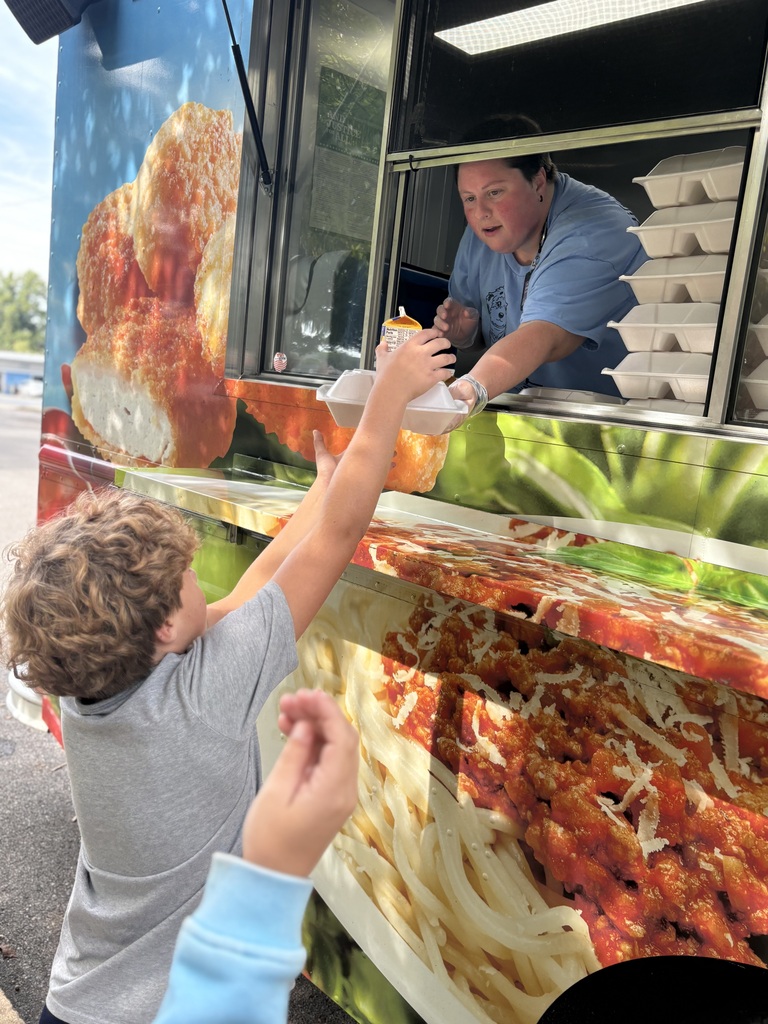 A student reaches up to receive an afternoon snack from a BTCS Nutrition Department staff member through the serving window of the colorful Munchbox food truck.