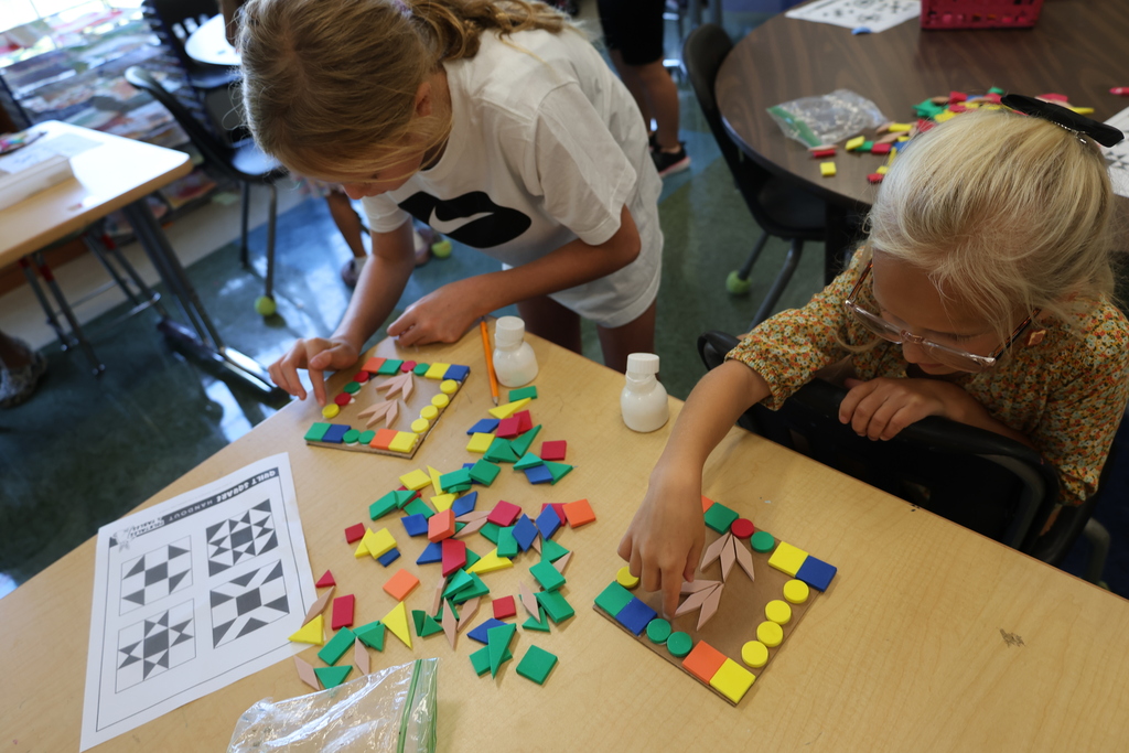 Two Fairmount Elementary students use colorful geometric shapes to create patterns and designs on cardboard squares during art class.