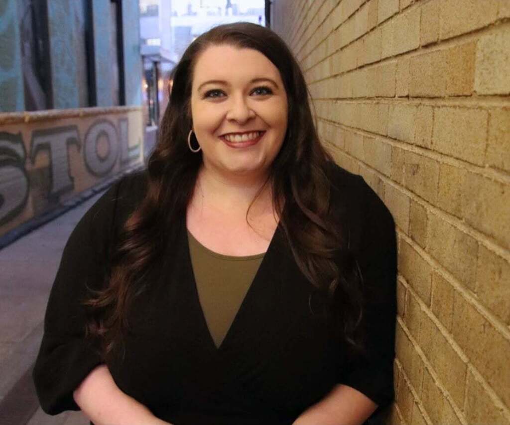 Autumn Roe smiles for a portrait while standing beside a tan brick wall. She has long dark hair and is wearing a black top.